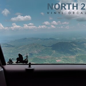 May include: A black vinyl decal of a person sitting by a campfire in the woods. The decal is on a car window with a view of mountains and clouds in the background. The text "NORTH 21 VINYL DECALS" is at the top of the image.