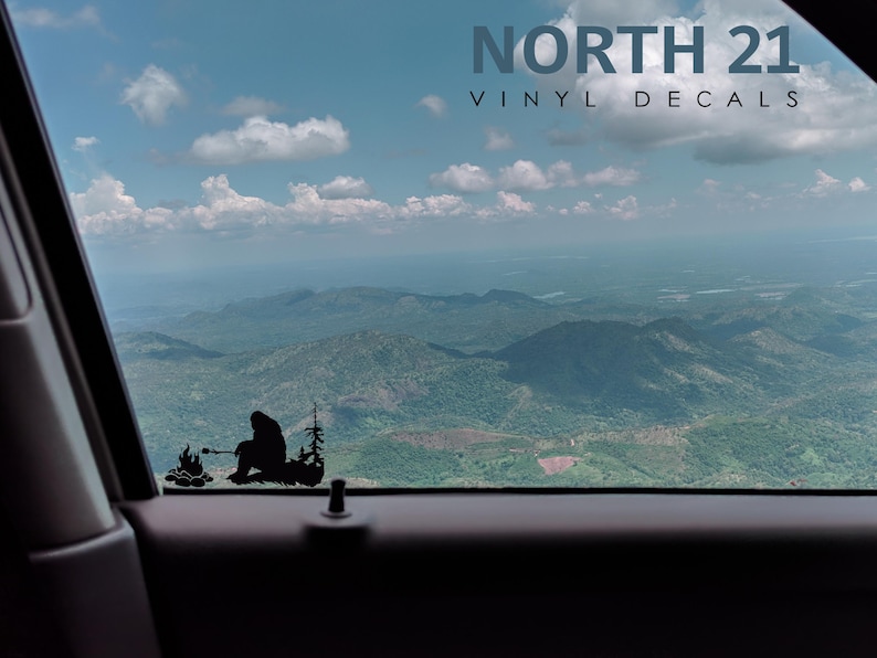 May include: A black vinyl decal of a person sitting by a campfire in the woods. The decal is on a car window with a view of mountains and clouds in the background. The text "NORTH 21 VINYL DECALS" is at the top of the image.