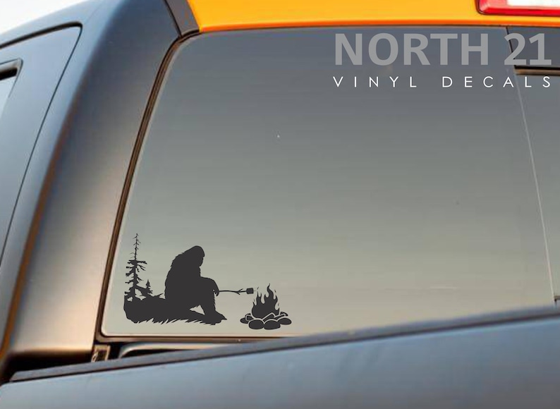May include: A black silhouette of a Bigfoot roasting a marshmallow over a campfire. The image is a vinyl decal for a car window. The text "NORTH 21 VINYL DECALS" is in the upper right corner of the image.