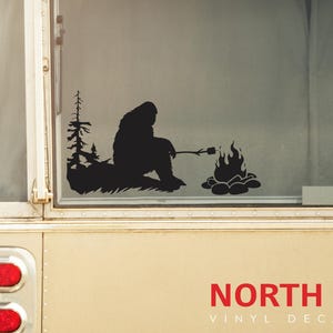 May include: Black vinyl decal of a Bigfoot silhouette roasting a marshmallow over a campfire. The decal is on a white camper van window. The text "NORTH 21 VINYL DECALS" is below the decal.