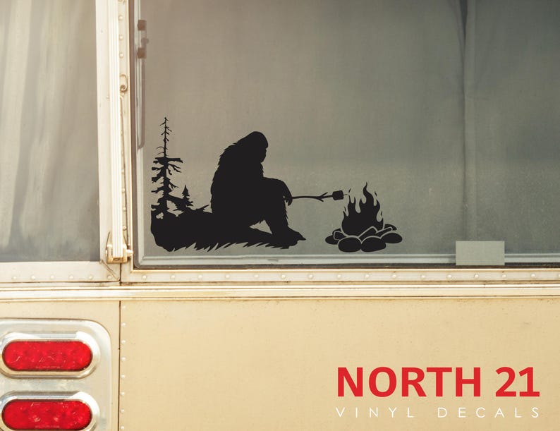 May include: Black vinyl decal of a Bigfoot silhouette roasting a marshmallow over a campfire. The decal is on a white camper van window. The text "NORTH 21 VINYL DECALS" is below the decal.
