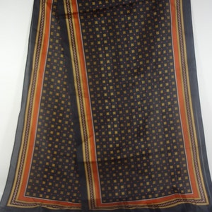 Op de afbeelding: Een zwarte zijden sjaal met een geometrisch patroon van kleine vierkanten en stippen in oranje en goud. De sjaal heeft een brede rand met strepen in oranje, goud en wit.