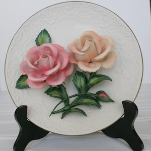 May include: A round white ceramic decorative plate featuring a raised, three-dimensional sculpture of two roses in the center. One rose is deep pink and the other is a soft peach color, both with green leaves and stems. The plate has a gold rim.