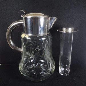 May include: A clear glass pitcher with a silver lid and a silver top for a bottle stopper. The pitcher has a cut glass design and a grape motif. The bottle stopper is a clear glass tube with a silver top.
