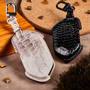 May include: Two leather keychains. One is off-white with a crocodile pattern, and the other is black with a similar texture. Both have metal clasps and are designed to hold keys. The keychains are displayed on a neutral surface.