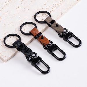 May include: Three keychains with black metal hardware and leather straps in black, brown, and olive green. Each keychain has a split ring and a lobster claw clasp for attaching keys or other items.