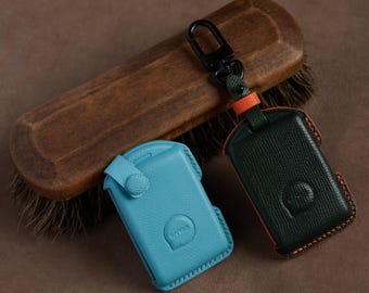 May include: Two textured leather key fob covers, one in bright turquoise and one in dark green with orange stitching. The dark green cover features a black metal swivel clasp attached by a small orange and green leather strap. Both cases have a circular embossed logo.