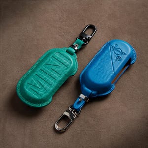 May include: Two textured leather key fob covers with metal clips. One cover is emerald green with the word MINI embossed on the surface. The other cover is bright blue and features the MINI logo embossed on the top section with a smooth finish.