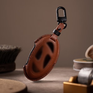 A brown leather keychain with a black metal clasp and a black leather loop is displayed on a wooden surface, surrounded by various objects.