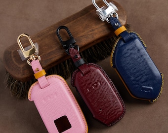 May include: Three textured leather car key fob covers in pink, dark red, and navy blue. Each cover features a metal swivel clasp, a small yellow leather accent loop, and contrast stitching. The pink cover has a square cutout on the front surface.