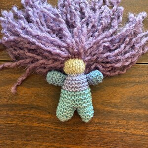 May include: A handmade doll with a light tan head, a body that fades from blue to purple, and blue arms. The doll has a large, fluffy, purple yarn hairpiece.