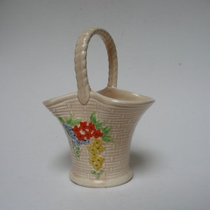 Puede incluir: Un pequeño jarrón de cerámica de color crema con forma de cesta con un patrón tejido y un asa. El jarrón está decorado con un diseño floral en tonos rojos, amarillos, azules y verdes.