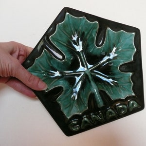 Puede incluir: Un azulejo de cerámica verde en forma de hoja de arce con la palabra "CANADA" escrita en él.