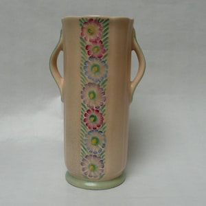 May include: A pale pink ceramic vase with two handles and a floral design. The vase has a green base and a row of pink, blue, and yellow flowers with green leaves.