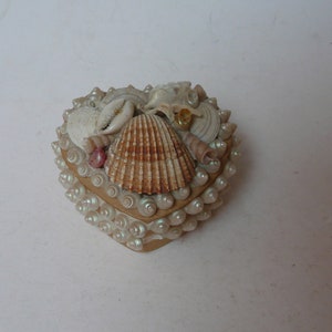 May include: A heart-shaped box made of seashells. The box is decorated with a variety of seashells, including a large, pink seashell in the center.