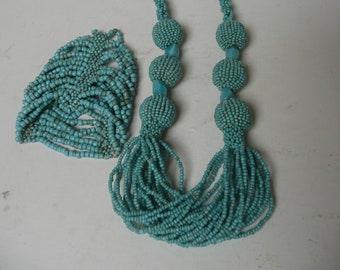 Puede incluir: Un conjunto de collar y pulsera de cuentas turquesas. El collar presenta grandes esferas de cuentas y largas borlas de cuentas. La pulsera es una banda ancha de cuentas turquesas. Las joyas se exhiben sobre una superficie blanca.