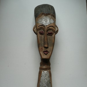 Puede incluir: Una escultura de madera de una cabeza humana con una banda de metal plateado en la frente. La escultura está pintada de marrón, rojo y negro. La escultura está sobre una base de madera.