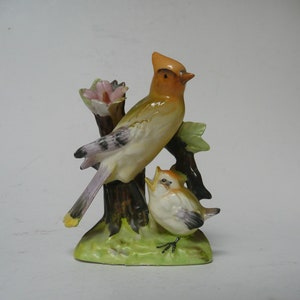 May include: A porcelain figurine of two birds perched on a branch. The larger bird is yellow, orange, and brown with a pink flower behind it. The smaller bird is white with brown accents. The base is green and brown.