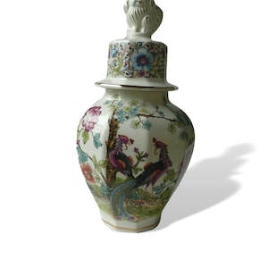 Peut inclure: Un vase en céramique blanche avec une bordure dorée et des motifs floraux. Le vase a un couvercle avec un petit lion en céramique blanche comme bouton. Le vase est décoré d'un motif floral et de deux paons.