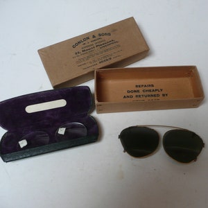 May include: A pair of vintage clip-on sunglasses with dark green lenses and a gold metal frame. The sunglasses are in a brown cardboard box with the text "REPAIRS DONE CHEAPLY AND RETURNED BY" printed on it. The sunglasses are also in a black leather case with a purple velvet lining.