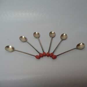 May include: Six silver teaspoons with red plastic handles arranged in a fan shape on a white surface.
