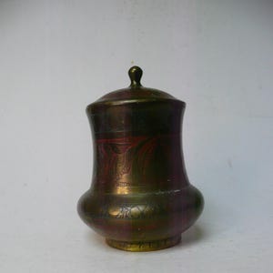 May include: A small, brass, decorative jar with a lid. The jar has an ornate design with red and gold accents.