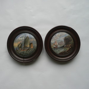 Puede incluir: Dos pinturas redondas enmarcadas de barcos en el mar. Las pinturas están en marcos marrones oscuros con un borde dorado. Las pinturas están sobre un fondo blanco.