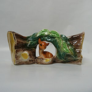May include: A ceramic planter shaped like a tree trunk with a deer peeking out from behind green leaves. The planter is brown with yellow accents and has a hole in the center for planting.