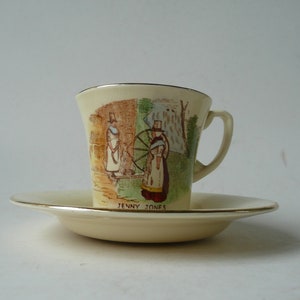 May include: A cream-coloured teacup and saucer set with a gold rim. The teacup features a vintage illustration of two women in traditional Welsh dress, one spinning a wheel. The saucer has the words "Jenny Jones" printed on the bottom.