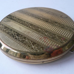 May include: A gold-colored compact mirror with a decorative pattern of stripes and floral designs.