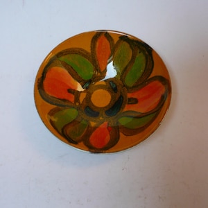 Könnte beinhalten: Kleine, runde Keramikschale mit floralem Design. Die Schale hat einen gelb-orangenen Hintergrund mit einer großen Blume in der Mitte. Die Blume hat rote, grüne und orangefarbene Blütenblätter mit schwarzen Umrissen. Die Schale dient zum Servieren kleiner Portionen.