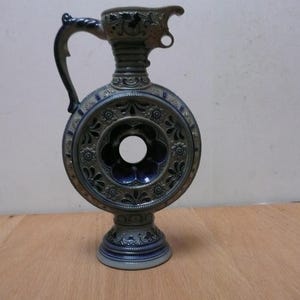 Puede incluir: Una jarra de cerámica circular con un orificio central, un cuello alto con pico vertedor y un asa curva. La pieza presenta un acabado en esmalte gris y azul profundo con intrincados patrones florales y geométricos en relieve por todo el cuerpo.