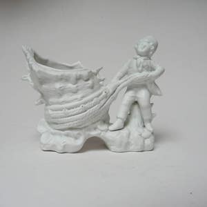 May include: White porcelain figurine of a boy pulling a rope attached to a large seashell. The seashell is shaped like a shoe and has a small opening at the top.