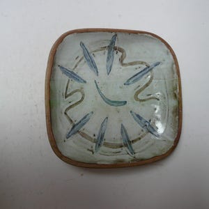 Op de afbeelding: Vierkant keramisch schaaltje met een lichtgroene en witte achtergrond. Het schaaltje heeft een bruine rand en heeft een ontwerp van blauwe bladachtige vormen en bruine gebogen lijnen. Het schaaltje is waarschijnlijk handgemaakt.