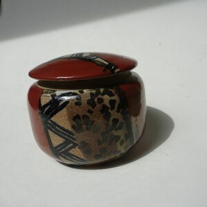 May include: A small, round ceramic jar with a lid. The jar is mainly red with black and tan abstract designs. The lid is red with a black and white design. The jar is likely handmade and suitable for storing small items.