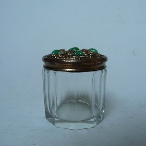 Könnte beinhalten: Ein klares Glasgefäß mit einem goldenen Deckel, der mit grünen Edelsteinen verziert ist. Das Gefäß hat eine sechseckige Form und ist perfekt zum Aufbewahren von kleinen Gegenständen.