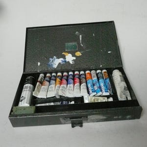 Puede incluir: Una caja de artista de metal gris oscuro abierta, llena de varios tubos de pintura. Los tubos están dispuestos en filas, mostrando una gama de colores que incluyen azul, rojo, amarillo y blanco. También se ve un recipiente de pintura negro.