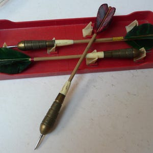 May include: A set of vintage darts with brass tips, wooden shafts, and feathered flights in a red plastic tray. The darts have green and burgundy coloured flights. The tray has moulded supports to hold the darts in place.