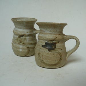 May include: Two handmade ceramic mugs with a speckled beige finish and a flared rim. Each mug features a rounded body with horizontal ridges and a dark brown painted abstract leaf or floral motif on the front. One mug has a curved handle.
