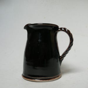 Puede incluir: Una jarra de cerámica marrón oscuro con un asa trenzada y un pico. La jarra tiene un acabado brillante y una base marrón claro. El diseño es simple y clásico, adecuado para servir bebidas.