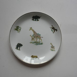 Puede incluir: Plato de cerámica blanco con borde dorado que presenta una variedad de animales, incluyendo una jirafa, un rinoceronte, una cebra, un elefante y un león. Los animales están representados en un estilo de dibujos animados y están rodeados de follaje verde.