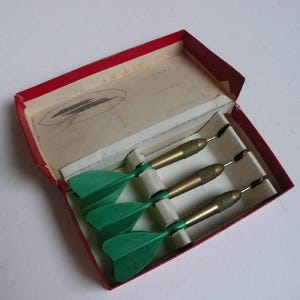 May include: A set of three darts with green flights and brass barrels, neatly arranged inside a red and cream-colored box. The box has a red border and a drawing on the inside lid. The darts are designed for target practice.