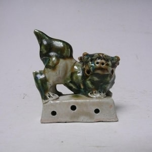 May include: A ceramic guardian lion figurine, featuring green and brown glaze details. The lion is posed atop a rectangular base with three holes. The figurine has a detailed mane and a raised tail.