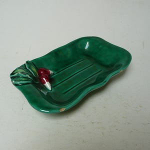 May include: A rectangular green ceramic soap dish with wavy edges and a glossy finish. The dish features a molded relief of a radish with red and white coloring on one side and three parallel drainage grooves running along the center base.