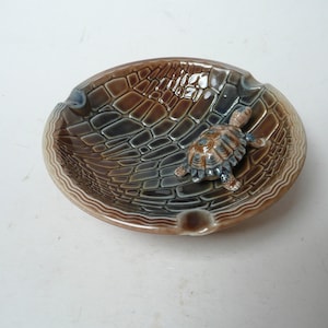 以下が含まれることがあります： 亀のデザインが施された茶色のセラミック製の灰皿。灰皿は亀の甲羅のような質感のある表面をしています。亀は茶色で、薄い茶色の甲羅をしています。