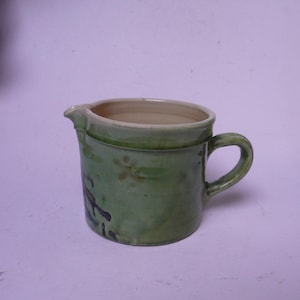 May include: A green ceramic pitcher with a white rim and a handle. The pitcher has a floral design and a brown glaze.