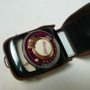 May include: Vintage black and burgundy light meter with a brown leather case. The meter has a circular dial with numbers and the name "Lunasix 2" printed on it. The case is open, revealing the meter's face.