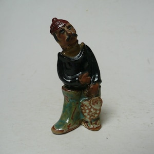 May include: A small ceramic figurine of a man wearing a red hat and black shirt. The figurine has a mustache and is holding a patterned bag. The boots are teal and brown.