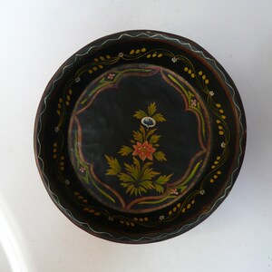 May include: A round wooden bowl with a black background and hand-painted floral designs in shades of green, yellow, pink, and white. The bowl has a scalloped edge and a white rim.
