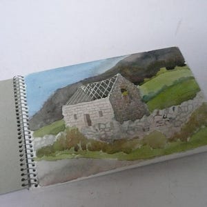 Puede incluir: Acuarela de un edificio de piedra con tejado parcialmente derrumbado, sobre un fondo de colinas verdes y una montaña oscura. La obra de arte está en un cuaderno de espiral, con un cielo azul claro.